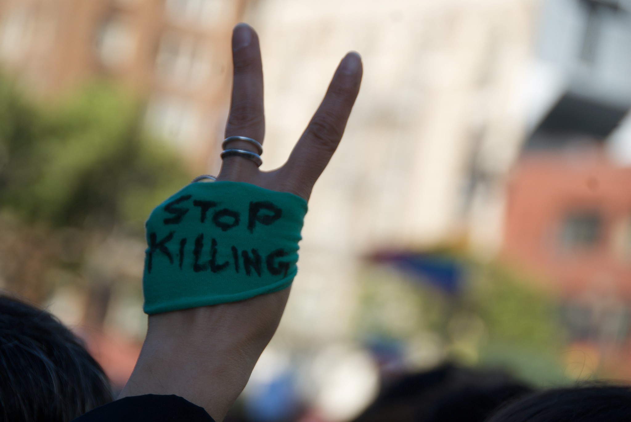 Green bandage that says “Stop Killing” wrapped around hand holding up a Peace sign.