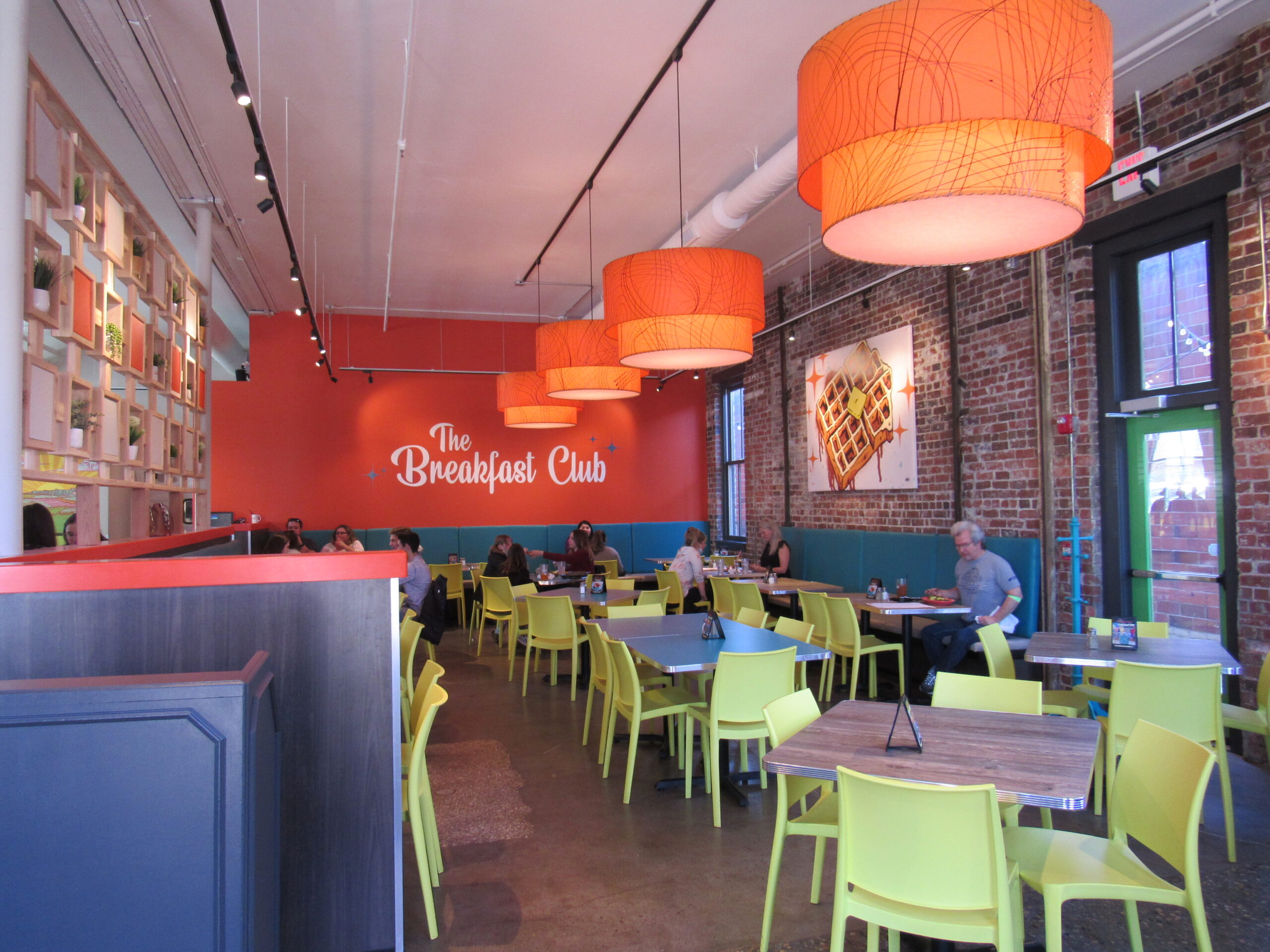 front view of dining room, orange back wall with “The Breakfast Club” written, customers eating at tables.
