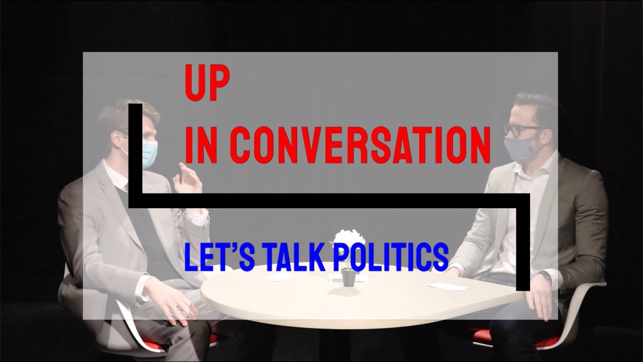 Kody and Matt sit at a table talking. A transparent logo for “UP in Conversation: Let’s Talk Politics” overlays the image.