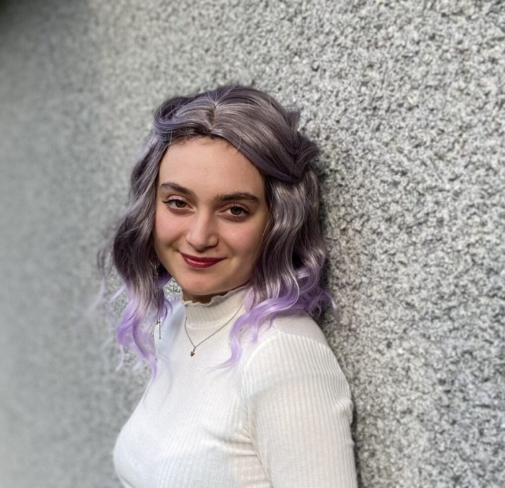 A picture of Agam with purple hair and wearing a white shirt leaning against a stone wall