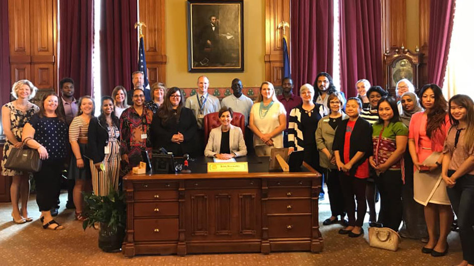 Governor Kim Reynolds poses for a photo at her desk, surrounded by a group of people