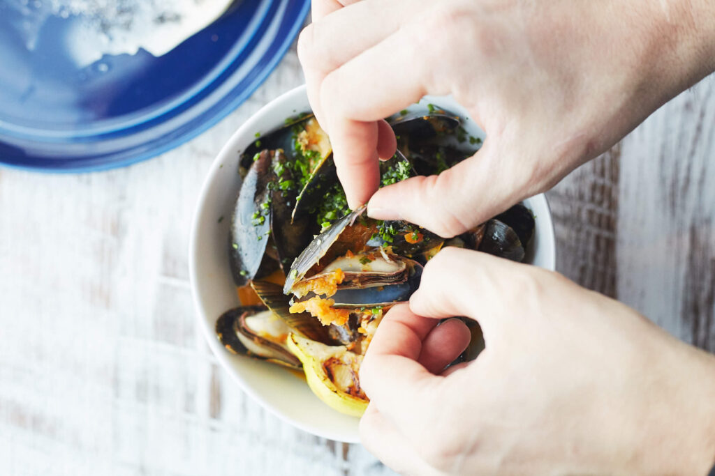 an up close shot of a hands opening a mussel above a bowl of cooked mussels