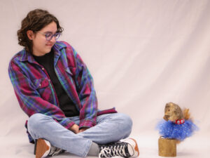 Julia, sitting on the floor in front of a white background, leaning away from a squirrel wearing a blue tutu mounted on a block of wood.