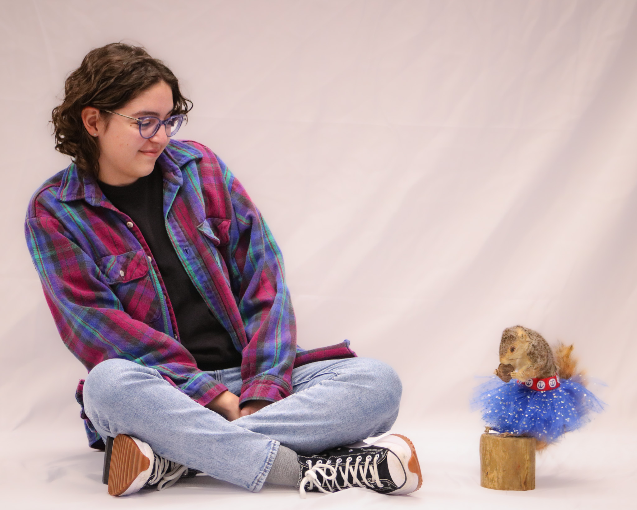 Julia, sitting on the floor in front of a white background, leaning away from a squirrel wearing a blue tutu mounted on a block of wood.