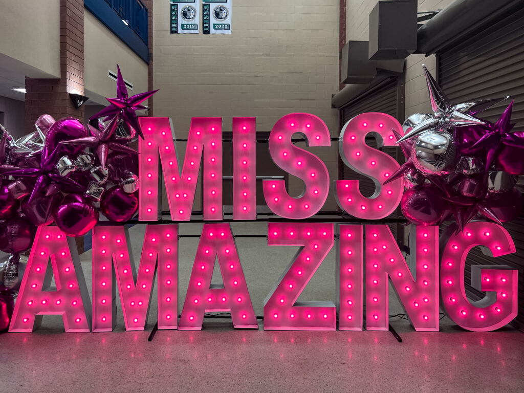 A pink sign that says “Miss Amazing” sits outside a highschool auditorium.
