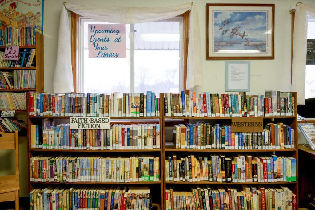 Two half bookshelves sit below a window that looks out onto Main Street of Garden Grove. They are full of books and labeled as “Faith-Based Fiction” and “Westerns." Next to the window, a painting of flying geese hangs on the wall.