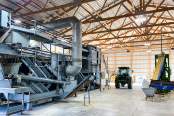 A large, metal system of tubes and conveyor belts is placed next to a tractor and sorting machine in a barn at Christensen Hop Farm.