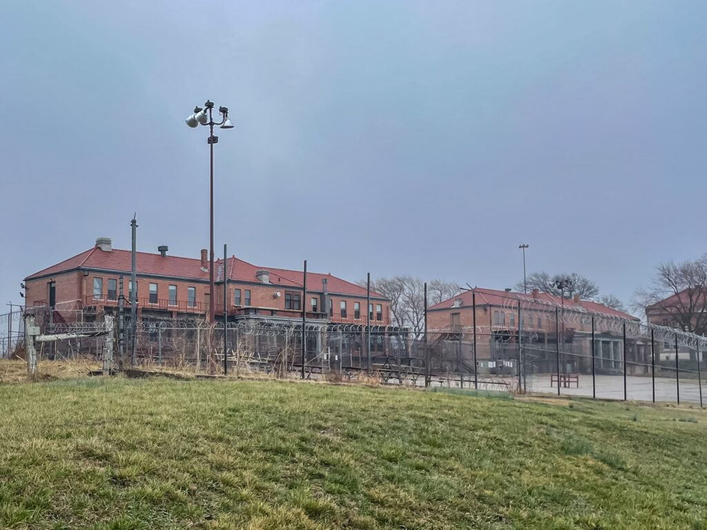 A chain link fence with barbed wire on top surrounds three red brick buildings. 