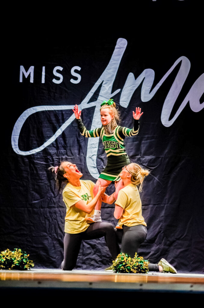 A little girl is lifted up by two volunteers in a cheerleading routine on stage.