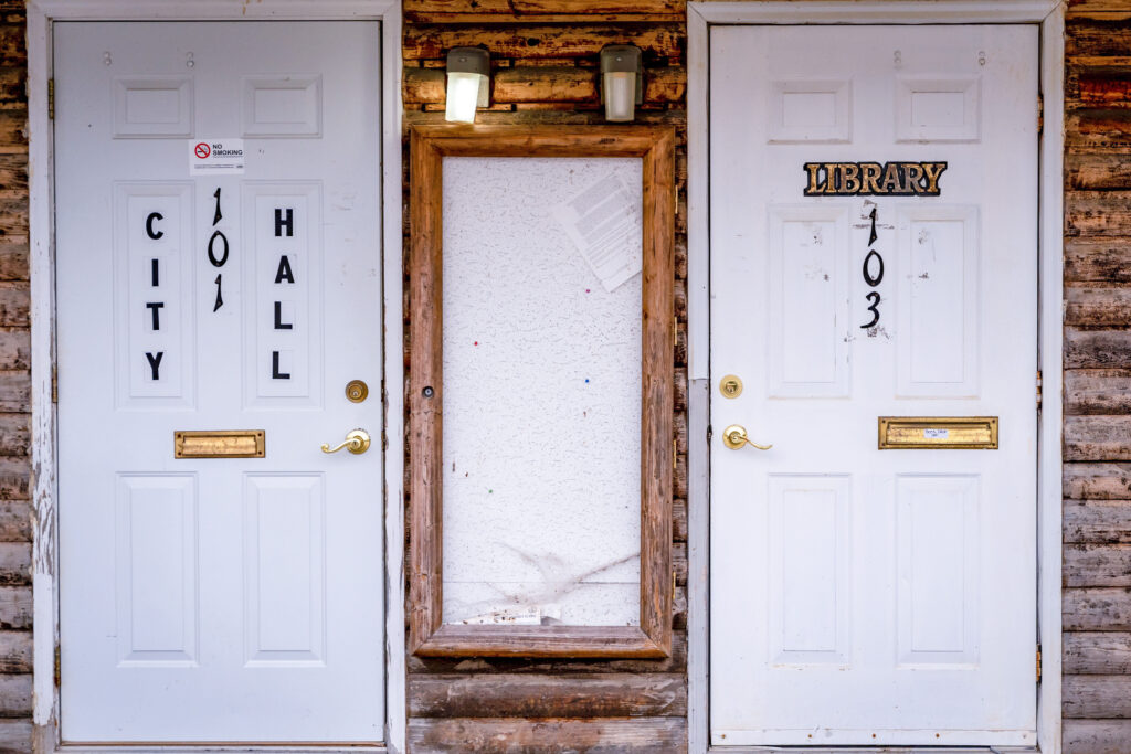 Two white exterior metal doors sit a couple of feet apart. The one on the left is labeled as “city hall” and the one on the right is labeled as “library.” In between the doors is a billboard with a single sheet of paper hanging crookedly.