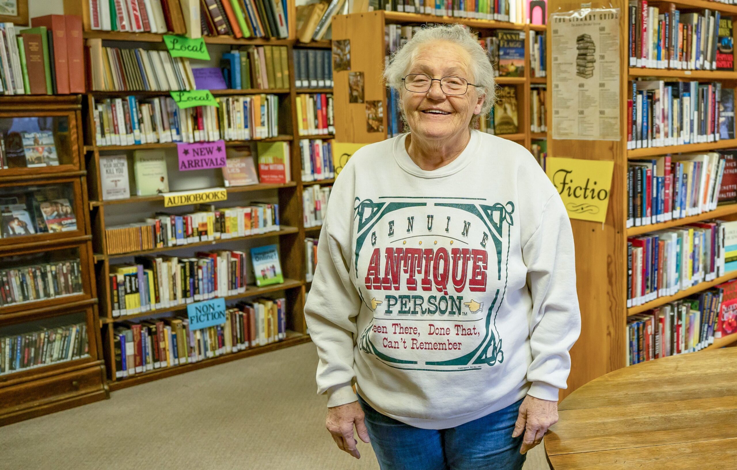 A woman with glasses and white hair stands in front of bookshelves. She’s wearing a gray crewneck sweatshirt that reads “Genuine Antique Person. Been There, Done That, Can’t Remember.” and jeans.