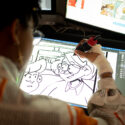 A person with short hair sits at a desk, drawing on a digital notepad. Steenz is drawing two cartoon figures on their notepad.