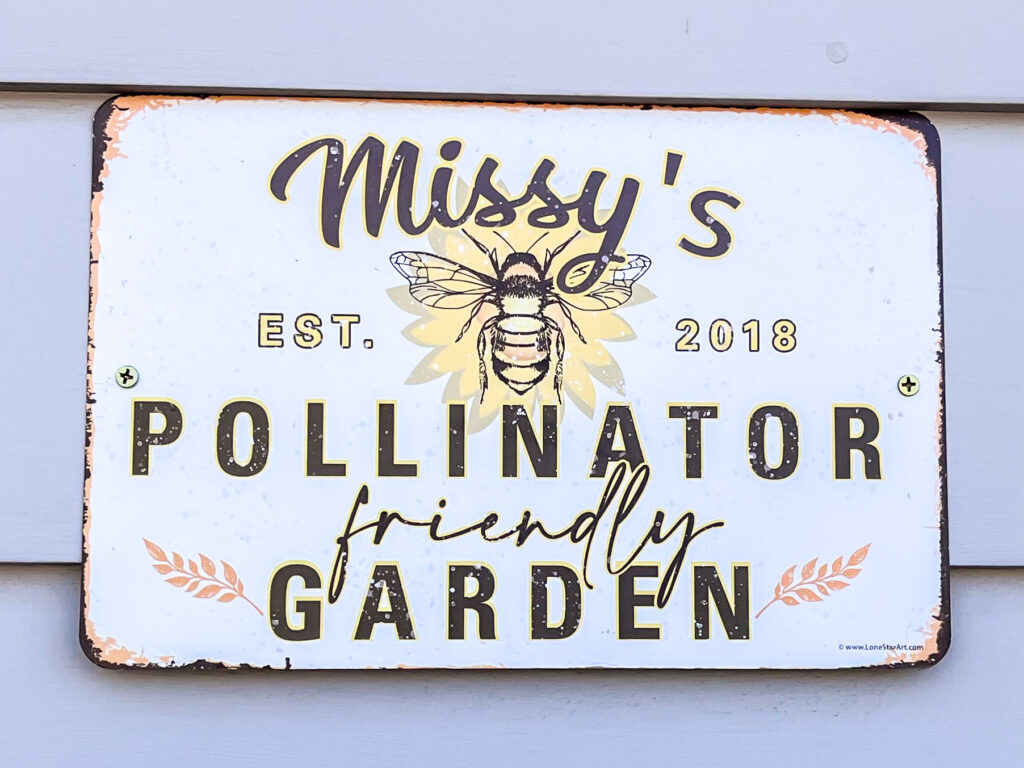 A rounded square sign reads “Missy’s pollinator friendly garden est. 2018” in black lettering with a yellow outline. In the top middle of the sign is a drawing of a bee on a light yellow flower. The corners of the sign have begun to rust.
