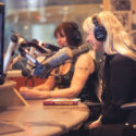 Two women, Lynette Reini-Grandell (front) and Venus de Mars (back), wearing headphones smile as they speak into professional microphones.