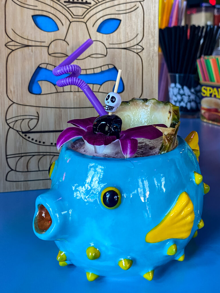 A purple drink served in a fish-themed cup garnished with a pineapple wedge and hibiscus flower. 
