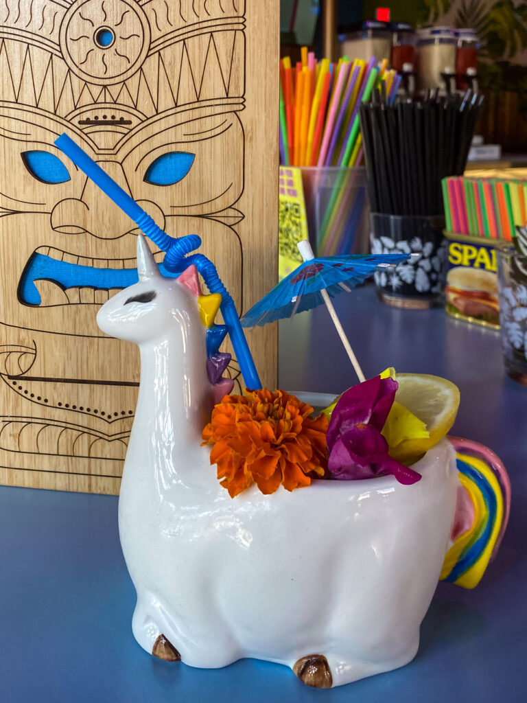  A clear drink served in a unicorn-themed cup garnished with a lemon slice and colorful flowers. 