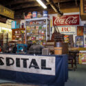 An image with various signs hanging on a wall, cabinets filled with antiques and a blue display table with a sign that says “Hospital” on the front.