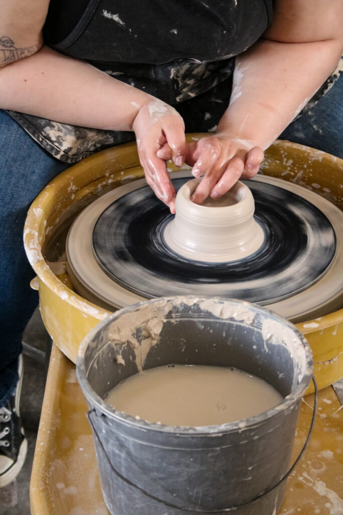 Urban Plains staff member, Kathryn, sits in front of the pottery wheel working on a bowl-like project.