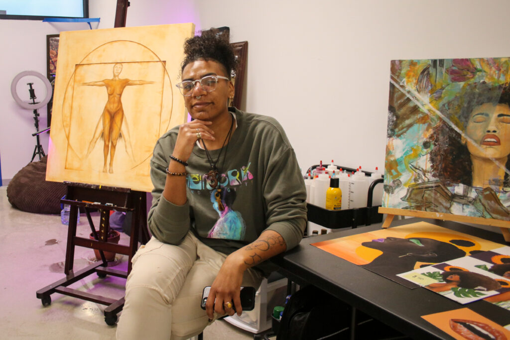 Artist Indigo Moore sits and poses in front of her art in her studio.
