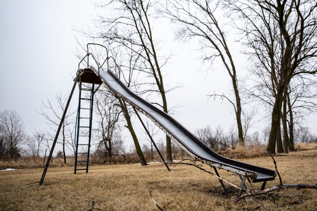 A worn metal slide lays in an abandoned park. 
