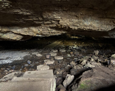 This image shows the inside of the cave. It is dark, but there is some dim lighting. The overhead rocks of the cave show the damp condition and the path of the cave has pools of water to wade through. There is both a cement path and rocks to step on to while walking through the cave. The rocks near the entrance have green moss on them.