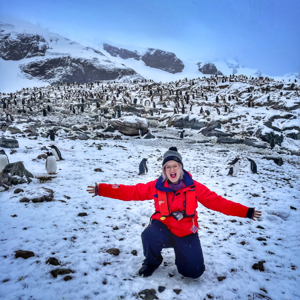 Donning a bright red coat and blue pants, Chelsea Hottovy poses for a photo in front of hundreds of penguins on a vast Antarctic landscape. 
