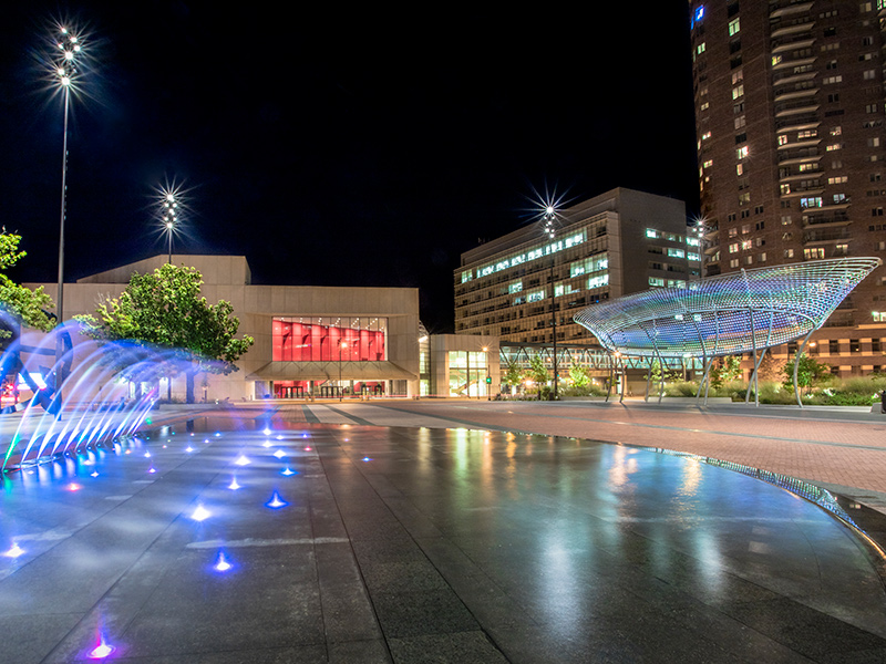 A large outdoor space at night just outside the Des Moines Civic Center. The park holds a large umbrella like sculpture and a fountain of water. 
