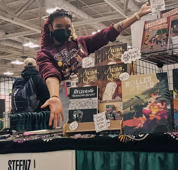 Steenz is standing at a convention showing off their work. They have several books on display including Archival Quality, Receiving Transmissions, Heart of the City and more.
