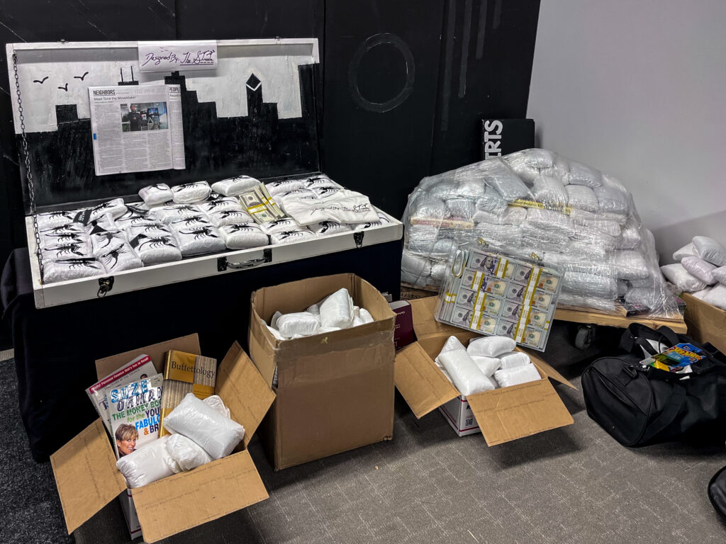 Several white packages with the image of a scorpion on them representing kilos of cocaine sit in an open box in Tone the MoveMaker’s studio. Surrounding the box are more packages in cardboard boxes and in a large stack wrapped in plastic wrap.
