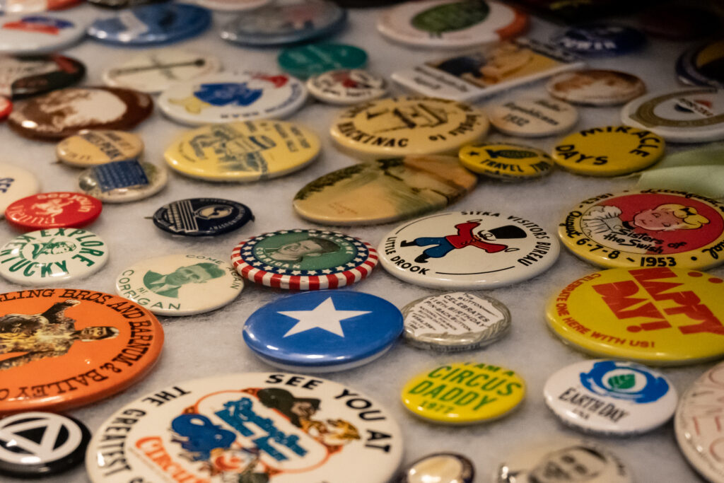 Close up of various buttons on a white background. Notable buttons in the foreground include: a blue button with a white star and a yellow button that says “circus daddy 1977” in green writing.
