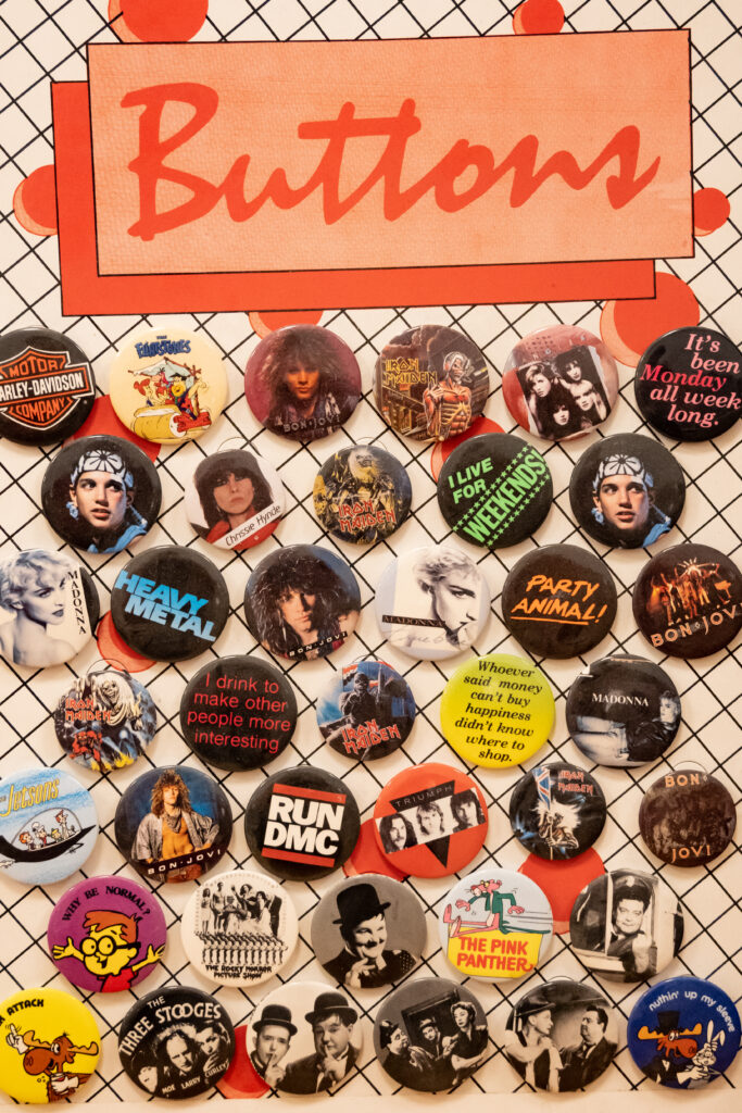 At the top in an 80s-style orange font the word “buttons.” Below that a variety of buttons covering popular 80s bands, comics, black and white movies, and more.

