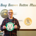 Co-Owner Joel Carter smiles, holding a plate-sized button with Jimmy Carter’s face on it and the inscription “Jimmy Carter for President in ‘76.” On the wall behind him it reads “Busy Beaver Button Museum.”