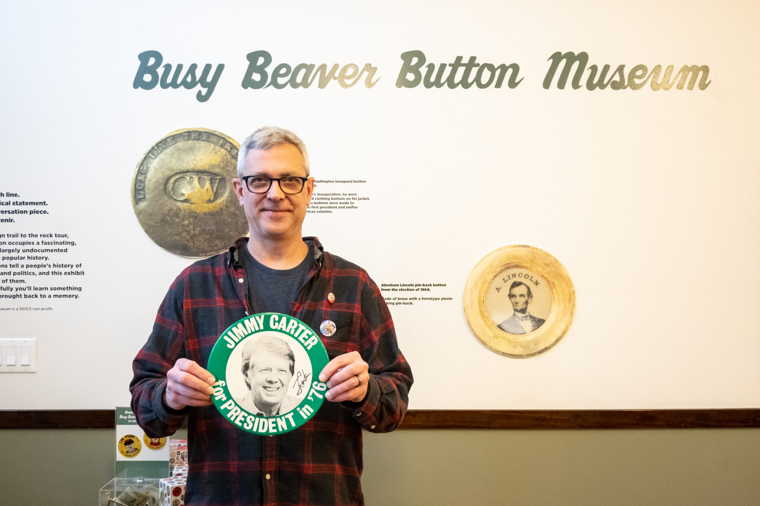 Co-Owner Joel Carter smiles, holding a plate-sized button with Jimmy Carter’s face on it and the inscription “Jimmy Carter for President in ‘76.” On the wall behind him it reads “Busy Beaver Button Museum.”