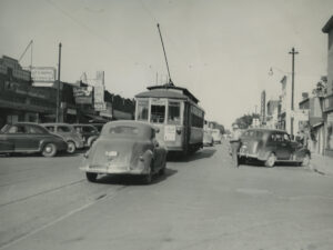 A historic photo shows a trolley on rails going down the street. A 1940s car follows behind. Several other cars are parked along the side of the road.