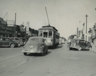 A historic photo shows a trolley on rails going down the street. A 1940s car follows behind. Several other cars are parked along the side of the road.